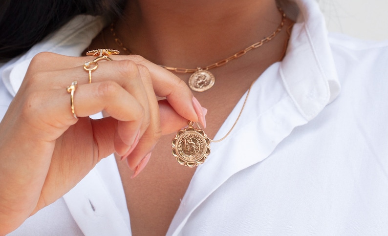 Peut inclure: Un collier en cha&icirc;ne dor&eacute;e avec deux pendentifs en forme de pi&egrave;ce de monnaie. Le plus grand pendentif pr&eacute;sente un design d&eacute;taill&eacute; et le plus petit pendentif est un simple cercle. Le collier est port&eacute; par une personne avec une chemise blanche.