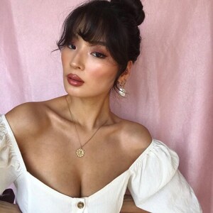 Peut inclure: Une femme aux cheveux fonc&eacute;s coiff&eacute;s en chignon avec une frange, portant un haut blanc &agrave; &eacute;paules d&eacute;nud&eacute;es avec un collier en or et des boucles d'oreilles en perles. Elle regarde la cam&eacute;ra.