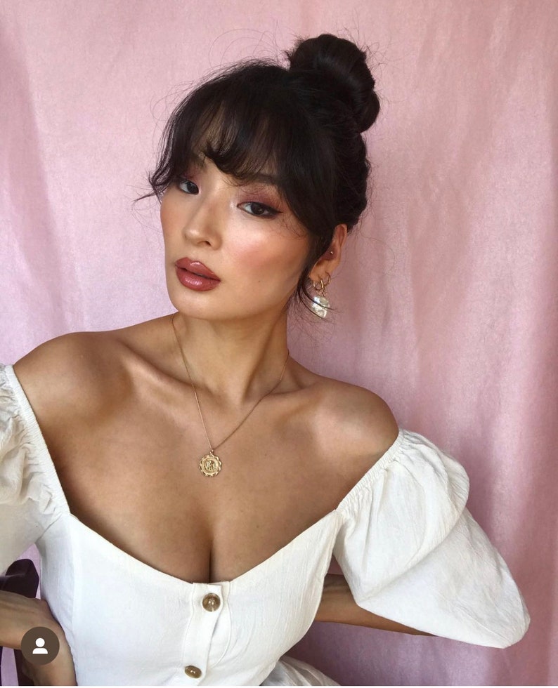 Peut inclure: Une femme aux cheveux fonc&eacute;s coiff&eacute;s en chignon avec une frange, portant un haut blanc &agrave; &eacute;paules d&eacute;nud&eacute;es avec un collier en or et des boucles d'oreilles en perles. Elle regarde la cam&eacute;ra.