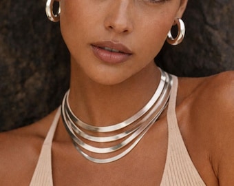 Op de afbeelding: Een zilverkleurige sieradenset bestaande uit een meerstrengs choker-ketting met vier gladde, platte, gebogen banden en een paar bijpassende dikke, gepolijste zilverkleurige oorringen. De sieraden hebben een reflecterende, metalen afwerking.