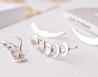 May include: A pair of silver ear climbers featuring a moon phase design, showing the transition from a full circle to a crescent shape. The earrings have a curved wire backing for secure placement along the earlobe, crafted in a polished silver finish.