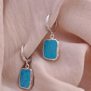 May include: A pair of silver hoop earrings featuring rectangular, rough-cut turquoise stone pendants. Each earring consists of a polished silver hoop connected to a vibrant blue, flat-faced turquoise slab with a textured, metallic silver edge.