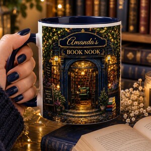 May include: A white ceramic mug with a dark blue interior and handle, featuring a detailed graphic of a cozy bookstore entrance illuminated by warm fairy lights. The text on the mug reads Amanda's BOOK NOOK, with a small sign saying Come in Get lost in a good book.