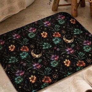 Può includere: Un tappetino da bagno rettangolare con sfondo nero e un design floreale e celeste. Il motivo include fiori colorati, lune crescenti e stelle. Il tappetino è posizionato su un pavimento piastrellato vicino a una vasca da bagno e pantofole.