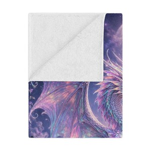 May include: A soft, white blanket with a detailed illustration of a fantasy dragon. The dragon features iridescent wings in shades of purple, pink, and blue, set against a starry night sky. The blanket is folded over, revealing a white, plush underside.
