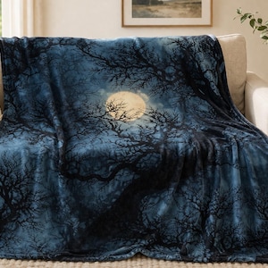 Puede incluir: Una manta azul oscuro con un diseño de bosque y luna, sobre un sofá beige. La manta representa árboles desnudos contra una luna llena. Hay un cojín decorativo en el sofá.
