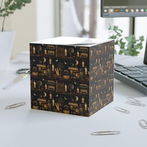 May include: A cube-shaped notepad with a repeating pattern of bookshelves, candles, globes, and moons in shades of gold and black. The top of the notepad is white, providing a contrast to the detailed design. The notepad is on a white surface.