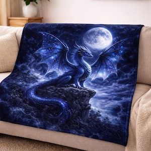 Könnte beinhalten: Eine blaue Drachendecke mit Fantasy-Motiv. Der Drache sitzt auf einem Felsen, die Flügel ausgebreitet, unter einem Vollmond. Die Decke ist über ein beiges Sofa drapiert.