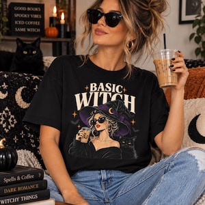 Può includere: Una t-shirt nera con la scritta BASIC WITCH in carattere color crema. La grafica raffigura una persona con un cappello da strega nero bordato di viola e occhiali da sole, che tiene in mano un caffè, accompagnata da un gatto nero.