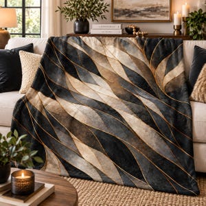 May include: A soft throw blanket featuring an abstract pattern of overlapping curved shapes in shades of charcoal gray, beige, and cream, accented by thin, metallic-toned gold lines that create a flowing, layered effect across the fabric surface.