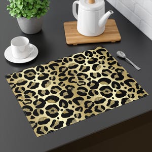 May include: A rectangular placemat with a gold and black leopard print pattern. The placemat is on a dark surface, with a white teacup and saucer, a teapot, and a small plant in the background.