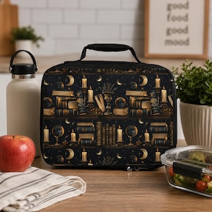 Op de afbeelding: Een rechthoekige lunchtas met een donker patroon van gestapelde boeken, brandende kaarsen, globes en maansikkels in goud en zwart. De tas heeft een zwart handvat aan de bovenkant en een ritssluiting met een gedetailleerde, thematische print.
