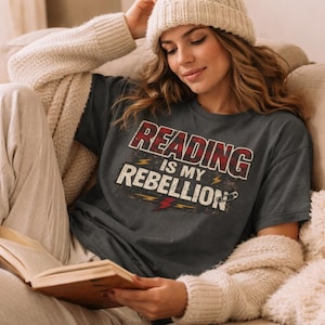 May include: Dark gray t-shirt with the text "READING IS MY REBELLION" in red and white lettering. The shirt has a vintage, distressed design. The person in the image is wearing a cream-colored beanie and sweater, and is holding a book.