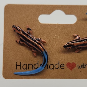 May include: A pair of lizard-shaped earrings attached to a brown cardboard display card. The lizards feature black and orange striped bodies with bright blue tails. The text "Handmade with love" with a red heart icon is printed on the card.