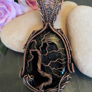 Könnte beinhalten: Ein Anhänger aus Kupferdraht mit einem schwarzen Achat-Edelstein, der mit einem Lebensbaum-Design verziert ist.