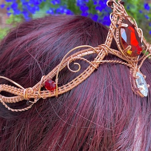 Peut inclure: Une couronne en fil de cuivre avec de grosses pierres précieuses rouges et transparentes. Le diadème artisanal présente un filigrane complexe et est orné d'une gemme rouge centrale en forme de goutte et de petits accents rouges et transparents.