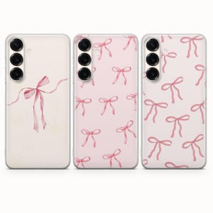 Pode incluir: Três capas de telemóvel com um design de laço rosa. A primeira capa tem um único laço, enquanto as outras duas apresentam um padrão repetido de laços. As capas têm uma borda transparente e um conjunto de câmeras.