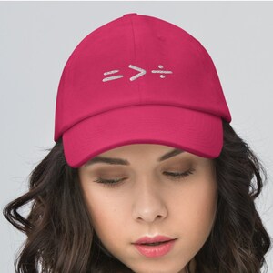 May include: A magenta baseball cap featuring embroidered white mathematical symbols: equals, greater than, and division. The cap is worn by a person wearing a white jacket.