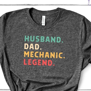 Puede incluir: Camiseta gris oscuro con las palabras "HUSBAND. DAD. MECHANIC. LEGEND." en verde azulado, amarillo y rojo. La camiseta est&aacute; anudada en la parte inferior. Las palabras "Deckorate Studios" est&aacute;n en la parte superior derecha.