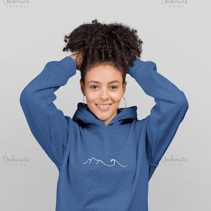 May include: A blue hoodie with a white mountain and wave design. The hoodie has a front pocket and a drawstring hood. The model is wearing hoop earrings and blue jeans.