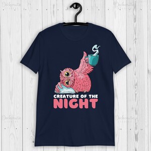May include: Navy blue t-shirt featuring a pink owl with a teal mug and the text "CREATURE OF THE NIGHT" in white and pink. The owl is holding a pillow. The shirt is hanging on a black hanger.