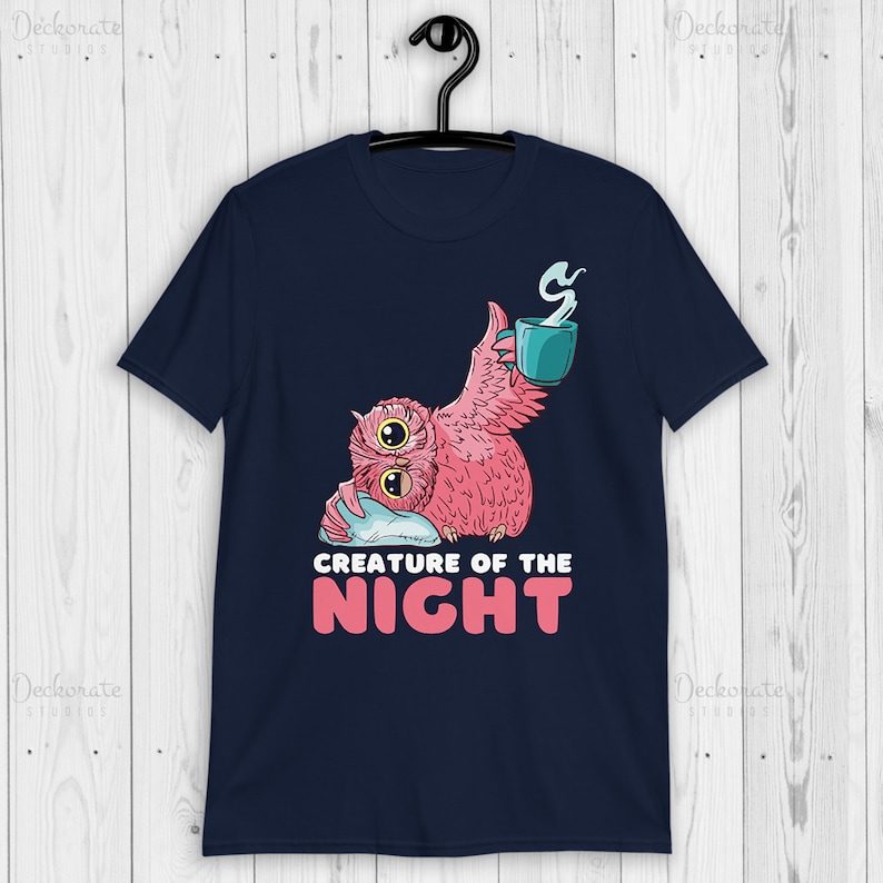 May include: Navy blue t-shirt featuring a pink owl with a teal mug and the text "CREATURE OF THE NIGHT" in white and pink. The owl is holding a pillow. The shirt is hanging on a black hanger.