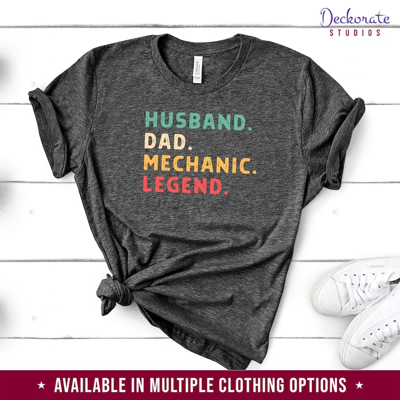Puede incluir: Camiseta gris oscuro con las palabras "HUSBAND. DAD. MECHANIC. LEGEND." en verde azulado, amarillo y rojo. La camiseta est&aacute; anudada en la parte inferior. Las palabras "Deckorate Studios" est&aacute;n en la parte superior derecha.