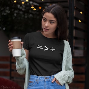 May include: Black t-shirt with a white graphic of mathematical symbols: equals, greater than, and division. The shirt is worn with blue jeans and a light green cardigan. A coffee cup is held in the left hand.