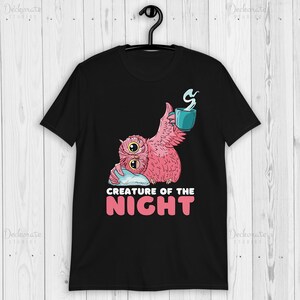 May include: Black t-shirt featuring a pink owl with two eyes, holding a teal mug with steam. The owl is resting on a pillow. The text "CREATURE OF THE NIGHT" is below the owl in white and pink.