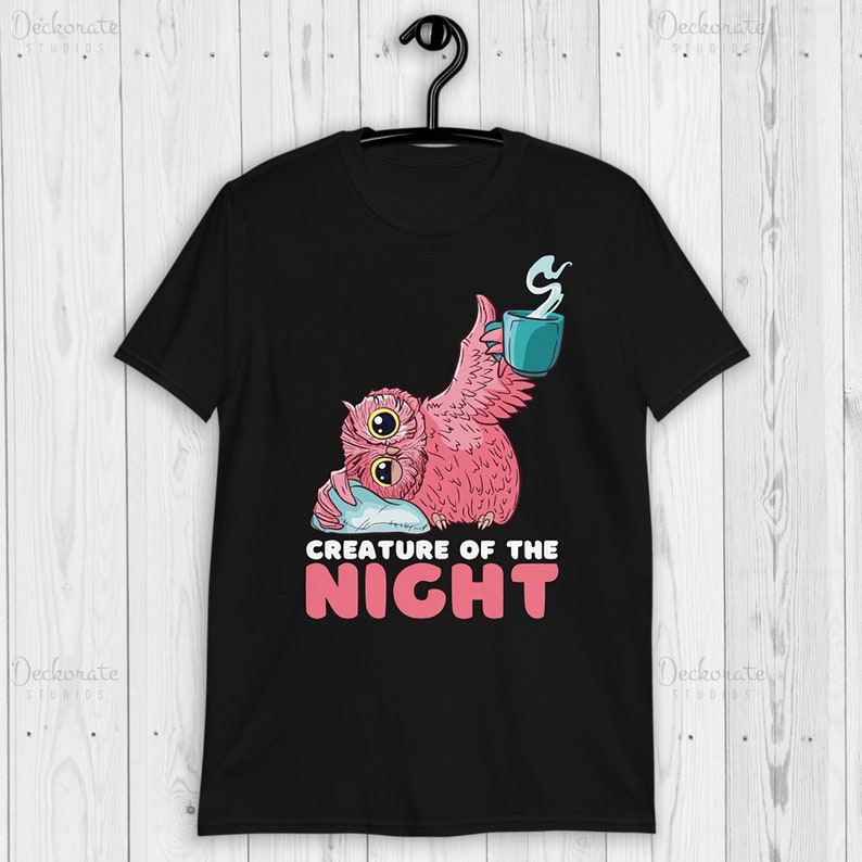 May include: Black t-shirt featuring a pink owl with two eyes, holding a teal mug with steam. The owl is resting on a pillow. The text "CREATURE OF THE NIGHT" is below the owl in white and pink.