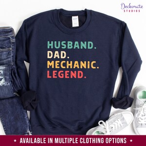 Puede incluir: Sudadera azul marino con las palabras "HUSBAND. DAD. MECHANIC. LEGEND." en verde azulado, amarillo y rojo. La sudadera se muestra con vaqueros y zapatillas.