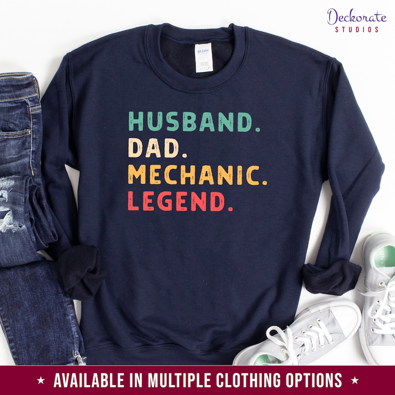 Puede incluir: Sudadera azul marino con las palabras "HUSBAND. DAD. MECHANIC. LEGEND." en verde azulado, amarillo y rojo. La sudadera se muestra con vaqueros y zapatillas.