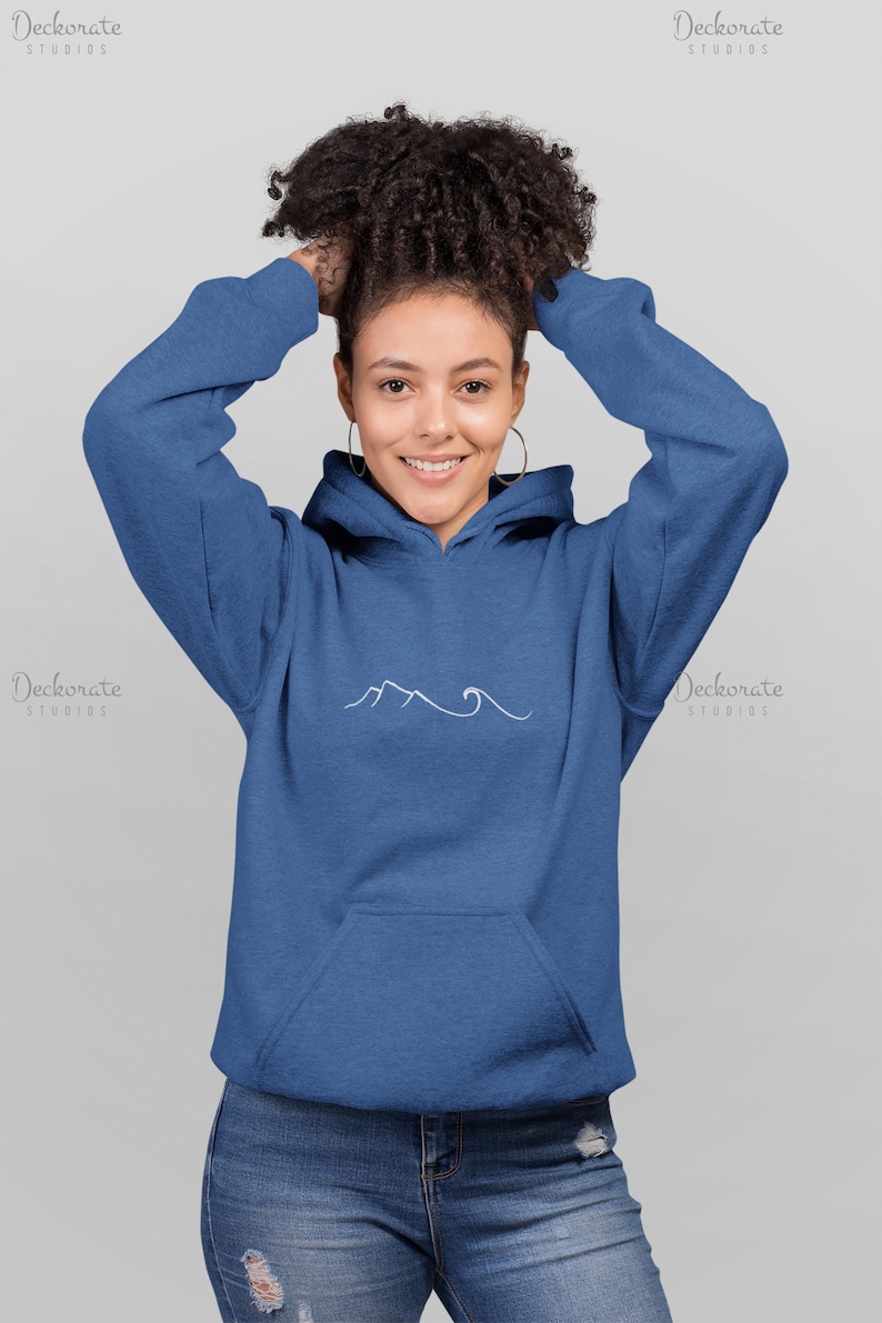 May include: A blue hoodie with a white mountain and wave design. The hoodie has a front pocket and a drawstring hood. The model is wearing hoop earrings and blue jeans.
