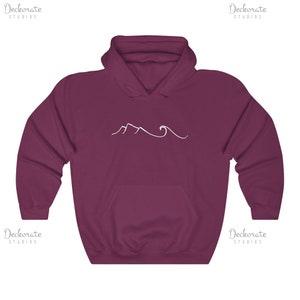 May include: Burgundy hoodie with a white mountain and wave design. The sweatshirt has a front pocket and a hood. The text "Deckorate Studios" is printed in white on the top right and bottom left corners.