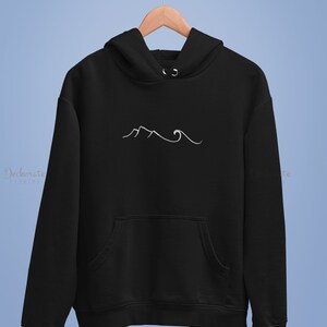 May include: Black hoodie with a white mountain and wave design. The hoodie has a front pocket and a hood. The design is minimalist and simple. The hoodie is hanging on a wooden hanger.