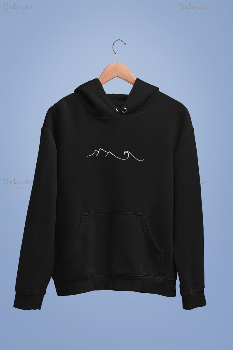May include: Black hoodie with a white mountain and wave design. The hoodie has a front pocket and a hood. The design is minimalist and simple. The hoodie is hanging on a wooden hanger.