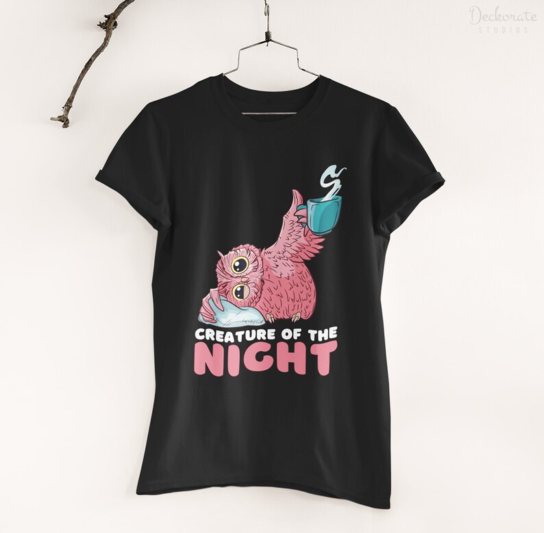 May include: Black t-shirt featuring a pink owl with a blue mug, resting on a pillow. The owl has large yellow eyes and is holding a mug with steam rising. The text "CREATURE OF THE NIGHT" is below the owl in pink letters.