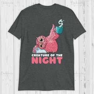 May include: Dark gray t-shirt featuring a pink owl with large eyes, holding a teal mug with steam rising. The text "CREATURE OF THE NIGHT" is below the owl in pink and white. The shirt is made of a soft material.