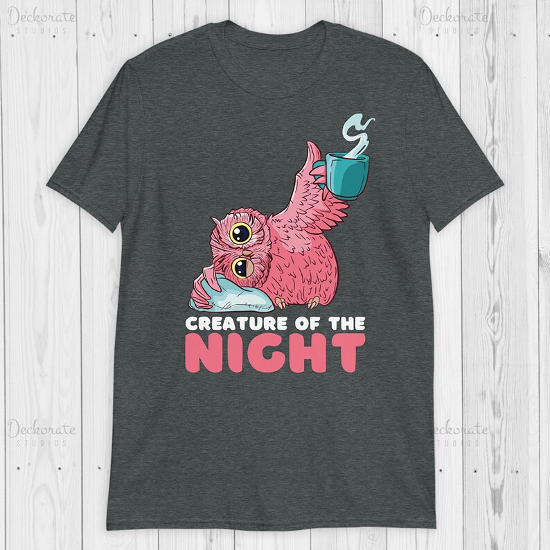 May include: Dark gray t-shirt featuring a pink owl with large eyes, holding a teal mug with steam rising. The text "CREATURE OF THE NIGHT" is below the owl in pink and white. The shirt is made of a soft material.