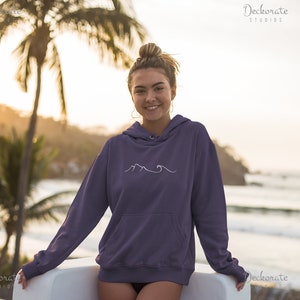 May include: A purple hoodie with a white mountain and wave design. The hoodie has a front pocket and a drawstring hood. The image shows a person wearing the hoodie, with a beach scene in the background.