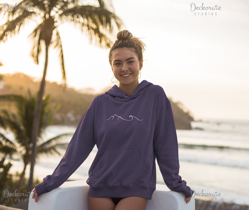 May include: A purple hoodie with a white mountain and wave design. The hoodie has a front pocket and a drawstring hood. The image shows a person wearing the hoodie, with a beach scene in the background.