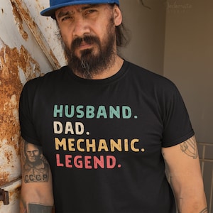 Puede incluir: Camiseta negra con las palabras "HUSBAND. DAD. MECHANIC. LEGEND." en letras turquesa, amarillo y rojo. La camiseta es de cuello redondo. La imagen tambi&eacute;n muestra a una persona con una gorra azul.