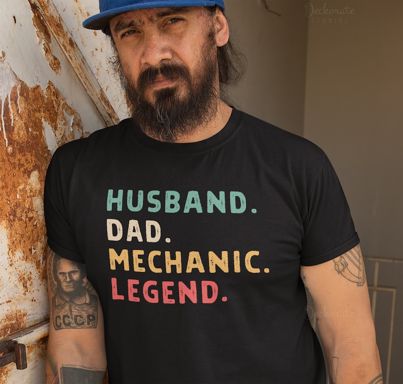 Puede incluir: Camiseta negra con las palabras "HUSBAND. DAD. MECHANIC. LEGEND." en letras turquesa, amarillo y rojo. La camiseta es de cuello redondo. La imagen tambi&eacute;n muestra a una persona con una gorra azul.