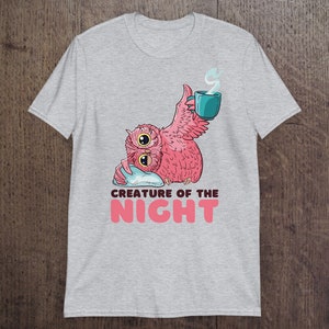 May include: A light gray t-shirt featuring a pink owl with two large eyes, holding a teal mug with steam rising. The text "CREATURE OF THE NIGHT" is below the owl in pink letters. The owl is resting on a blue pillow.