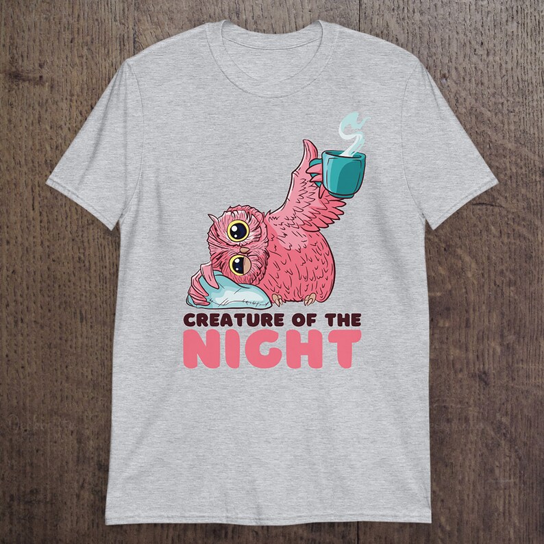 May include: A light gray t-shirt featuring a pink owl with two large eyes, holding a teal mug with steam rising. The text "CREATURE OF THE NIGHT" is below the owl in pink letters. The owl is resting on a blue pillow.