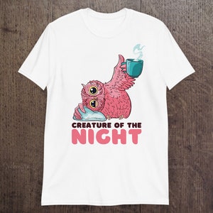 May include: White t-shirt featuring a pink owl-like creature with two large eyes, holding a teal mug with steam rising. The text "CREATURE OF THE NIGHT" is below the owl in pink letters. The shirt is displayed on a wooden surface.