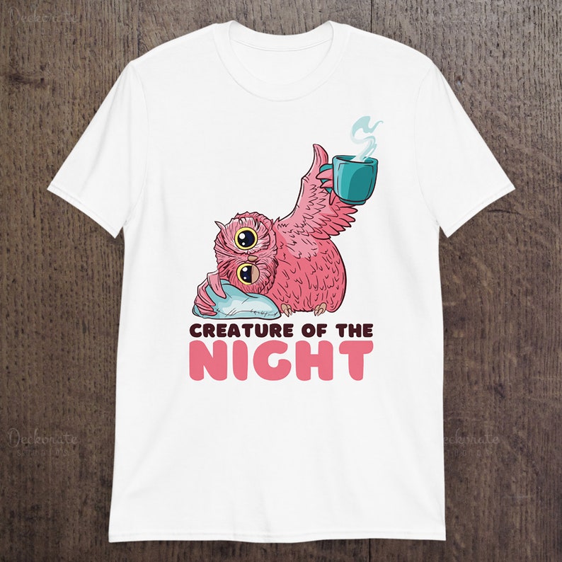 May include: White t-shirt featuring a pink owl-like creature with two large eyes, holding a teal mug with steam rising. The text "CREATURE OF THE NIGHT" is below the owl in pink letters. The shirt is displayed on a wooden surface.