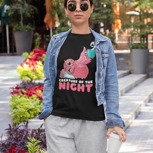 May include: Black t-shirt featuring a pink owl graphic with a teal mug and the text "CREATURE OF THE NIGHT" in white and pink. The owl is holding a pillow. The person is wearing a denim jacket and gray sweatpants.
