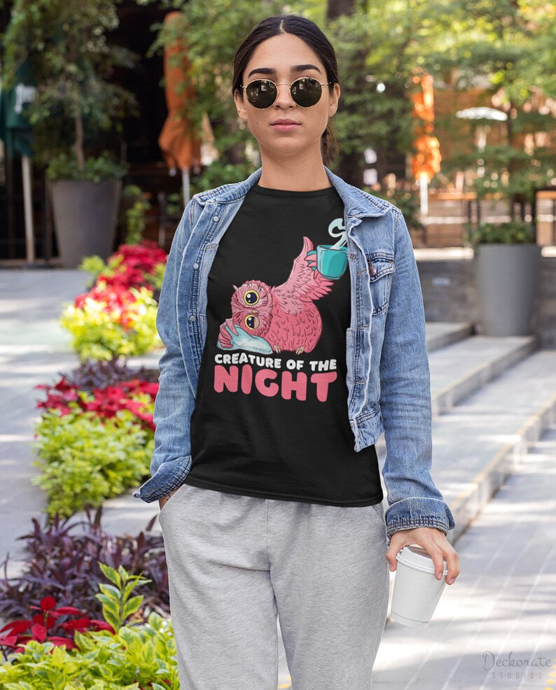 May include: Black t-shirt featuring a pink owl graphic with a teal mug and the text "CREATURE OF THE NIGHT" in white and pink. The owl is holding a pillow. The person is wearing a denim jacket and gray sweatpants.