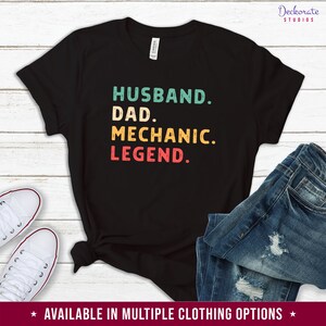 Puede incluir: Camiseta negra con las palabras "HUSBAND. DAD. MECHANIC. LEGEND." en verde azulado, amarillo y rojo. La camiseta se combina con vaqueros azules y zapatillas blancas. El texto "AVAILABLE IN MULTIPLE CLOTHING OPTIONS" est&aacute; en la parte inferior.